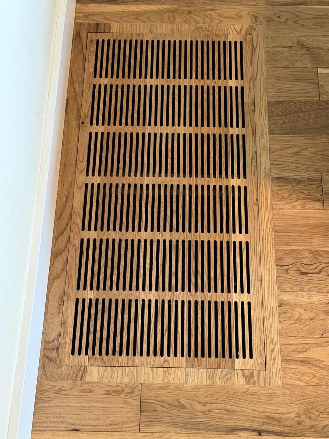 A large custom-milled oak return-air grille set flush into a white-oak floor beside the baseboard, its slot pattern carried across six panels